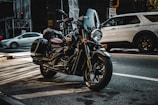 A black motorcycle is parked on a city street with its headlight turned on. The motorcycle is equipped with saddlebags and a windshield. In the background, a white SUV is visible along with blurred motion of another car. Urban elements such as a one-way street sign, traffic lights, and construction scaffolding are present.