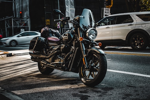 A black motorcycle is parked on a city street with its headlight turned on. The motorcycle is equipped with saddlebags and a windshield. In the background, a white SUV is visible along with blurred motion of another car. Urban elements such as a one-way street sign, traffic lights, and construction scaffolding are present.