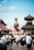 A crowd of people dressed in traditional attire participates in a cultural procession. They carry a large, ornately decorated bull statue through a street surrounded by traditional Balinese architecture. The sky is clear and blue, indicating a sunny day.