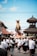 A crowd of people dressed in traditional attire participates in a cultural procession. They carry a large, ornately decorated bull statue through a street surrounded by traditional Balinese architecture. The sky is clear and blue, indicating a sunny day.