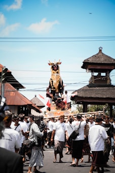 A crowd of people dressed in traditional attire participates in a cultural procession. They carry a large, ornately decorated bull statue through a street surrounded by traditional Balinese architecture. The sky is clear and blue, indicating a sunny day.