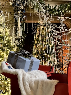 A festive scene with a red sleigh adorned with a soft, white fur throw. It holds a large, blue gift box tied with a shiny black ribbon. The background features decorated Christmas trees with twinkling lights, ornaments, and intricate, frosty branches. An ambiance of warmth and holiday cheer is created by the abundance of greenery and glowing decorations.