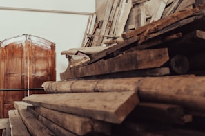 A stack of wooden planks and logs is piled on one side, suggesting materials for construction or woodworking. In the background, a large double wooden door is visible, along with some tools and additional planks leaning against a wall. The setting appears rustic and informal, likely a workshop or storage area for wood.