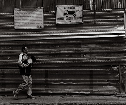 A person carrying a clock walks along a construction site with corrugated metal sheets. Two signs are visible on the wall, one in Spanish mentioning construction-related information and another warning about truck traffic.