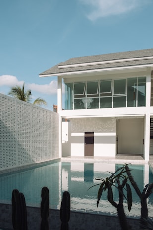A modern two-story house with a minimalist design is adjacent to a clear swimming pool. The structure features large glass windows and is surrounded by decorative walls with geometric patterns. Cacti are planted near the pool, and a palm tree is visible in the distance under a bright blue sky.