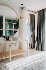 A modern bathroom with a large round mirror above dual sinks and decorative pendant lights hanging from the ceiling. The floor is covered in terrazzo tiles, and a window with sheer and dark curtains allows natural light to brighten the space. The walls feature green tiles, adding a touch of color and elegance.