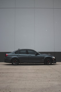 A sleek, dark-colored sedan is parked in front of a large, modern wall with clean lines and a muted color scheme. The car features tinted windows and black rims, adding to its stylish aesthetic.
