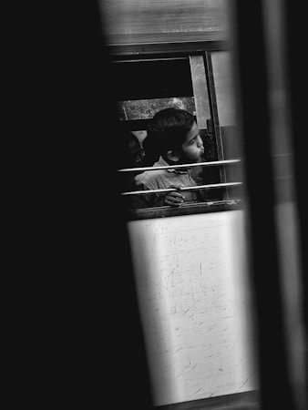 A young child gazes out from a bus window, framed by metal bars. The scene is captured in black and white, highlighting the child's expression and the surrounding textures. The bus interior is partially visible, with blurred elements in the foreground adding depth.
