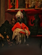 A collection of intricately detailed sculptures arranged in a display. The central figure is dressed in vibrant traditional attire with a turban and draped in orange and white fabric, adorned with jewelry. Surrounding it are other statues in dark hues, contributing to a rich, cultural aesthetic.