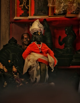 A collection of intricately detailed sculptures arranged in a display. The central figure is dressed in vibrant traditional attire with a turban and draped in orange and white fabric, adorned with jewelry. Surrounding it are other statues in dark hues, contributing to a rich, cultural aesthetic.