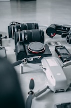 A collection of photography and videography equipment is arranged on a light surface. The items include several camera lenses, a compact camera with the brand name visible, a drone with partially visible propellers, memory cards, and a small action camera.