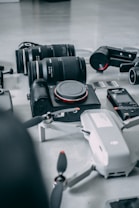 A collection of photography and videography equipment is arranged on a light surface. The items include several camera lenses, a compact camera with the brand name visible, a drone with partially visible propellers, memory cards, and a small action camera.