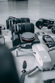 A collection of photography and videography equipment is arranged on a light surface. The items include several camera lenses, a compact camera with the brand name visible, a drone with partially visible propellers, memory cards, and a small action camera.
