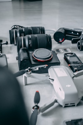 A collection of photography and videography equipment is arranged on a light surface. The items include several camera lenses, a compact camera with the brand name visible, a drone with partially visible propellers, memory cards, and a small action camera.