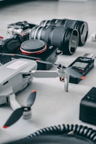 A collection of photography and videography equipment spread out on a flat surface, including a camera with interchangeable lenses, a drone, a voice recorder, and a GoPro.