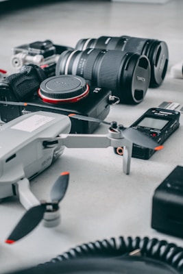 A collection of photography and videography equipment spread out on a flat surface, including a camera with interchangeable lenses, a drone, a voice recorder, and a GoPro.