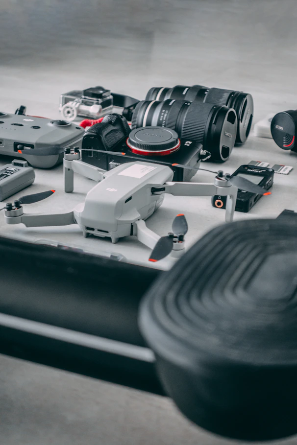 A collection of photography and videography equipment is arranged on a flat surface. The setup includes two camera lenses, a drone, a camera, and various camera accessories such as batteries, memory cards, and a remote controller. The equipment appears new and is positioned neatly, suggesting preparation for a professional shoot.
