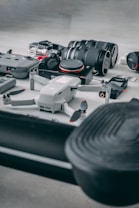 A collection of photography and videography equipment is arranged on a flat surface. The setup includes two camera lenses, a drone, a camera, and various camera accessories such as batteries, memory cards, and a remote controller. The equipment appears new and is positioned neatly, suggesting preparation for a professional shoot.