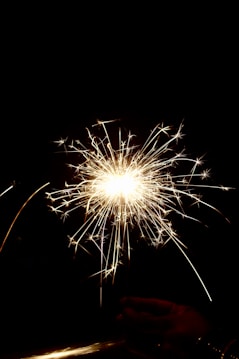 Bright, sparkling fireworks emit luminous trails of light in a burst pattern against a dark background, with a hand holding the sparkler at the bottom.