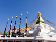 A large, impressive stupa with a white dome and golden spire stands against a clear blue sky. Colorful prayer flags are draped over the structure, with several poles, each topped with a golden ornament, lined up in front. The scene conveys a sense of peace and spirituality.