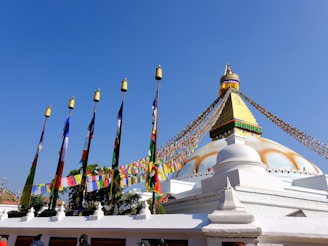 A large, impressive stupa with a white dome and golden spire stands against a clear blue sky. Colorful prayer flags are draped over the structure, with several poles, each topped with a golden ornament, lined up in front. The scene conveys a sense of peace and spirituality.
