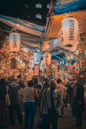 A bustling night market scene with a large crowd of people walking and interacting. Traditional Japanese lanterns with kanji characters hang overhead, casting warm light over the vibrant stalls. Many people wear face masks, and there are various colorful goods and decorations displayed throughout the market.