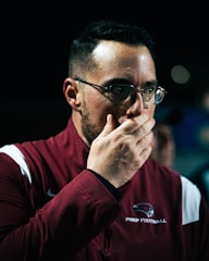 A man wearing glasses and a maroon sports jacket with a logo that reads 'Prep Football' is covering his mouth with his hand. The lighting is focused on his face, creating a serious and contemplative atmosphere.