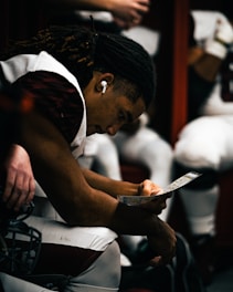 A football player with dreadlocks is seated in a dimly-lit locker room, wearing earphones and looking intently at a play chart. He appears focused and deep in thought as teammates are partially visible around him.