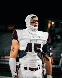 A football player is wearing a white uniform with the number 45 and the word 'PREP' on the chest area. The player is equipped with protective gear and has eye black under the eyes. The background is out of focus, possibly a stadium setting at night.