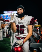 A football player in a white jersey with the number 16 and the word PREP on it smiles at the camera. He is holding a red football helmet and wearing a black headgear. Another hand is pointing at him from the left side of the image. Other players and a sports field with blurred lights are visible in the background.