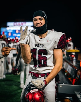 A football player in a white jersey with the number 16 and the word PREP on it smiles at the camera. He is holding a red football helmet and wearing a black headgear. Another hand is pointing at him from the left side of the image. Other players and a sports field with blurred lights are visible in the background.