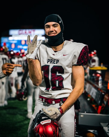 A football player in a white jersey with the number 16 and the word PREP on it smiles at the camera. He is holding a red football helmet and wearing a black headgear. Another hand is pointing at him from the left side of the image. Other players and a sports field with blurred lights are visible in the background.