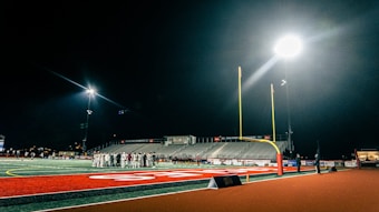 A dimly lit football field is observed at night with a group of people gathered near the center of the field. The stadium lights illuminate the field, creating a stark contrast against the dark sky. The goalpost stands tall in the foreground, and the empty bleachers are visible in the background.
