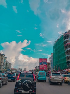 A bustling urban street with heavy traffic as several cars fill the lanes on a wide road. Buildings line both sides with a prominent construction site on the right, wrapped in green netting. A large billboard dominates the scene in the distance. The sky is bright with dramatic clouds, suggesting fair weather.