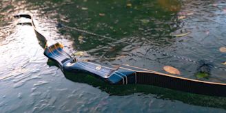 A sleek, modern recurve bow rests on a wet surface with scattered leaves. The bow has a glossy finish with a deep blue and black color, accented by fine lines and detailing. The string is taut, and the water reflects light, creating a serene and somewhat reflective atmosphere.