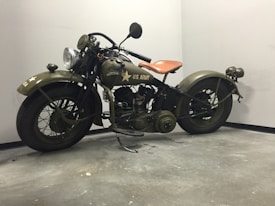 A vintage military motorcycle with olive green paint, featuring 'U.S. Army' markings and a star emblem. The bike has classic design elements such as a large headlamp, a tan leather seat, and robust tires. It is positioned against a plain, grey corner of a room, enhancing its historical and rugged appearance.