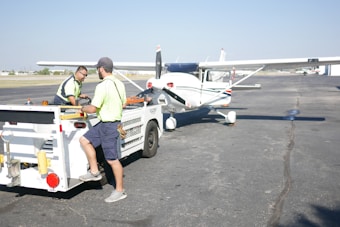 Two people are working on or near a small aircraft at an airport. One person is sitting on the back of a small vehicle with various equipment, while the other is standing nearby. Both are wearing high-visibility vests. The aircraft is a single-engine propeller plane parked on the tarmac under a clear blue sky.