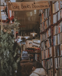 A cozy bookstore with shelves towering full of books. Two people are browsing the shelves, one wearing a light blue beanie and the other wearing a cream-colored jacket. There are festive red garlands decorating the shelves and a sign overhead that reads 'MORE BOOKS DOWNSTAIRS'. Warm lighting creates a welcoming atmosphere.