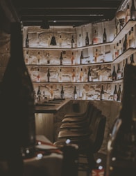 A dimly lit wine cellar with numerous wine bottles placed on wooden shelves that span across the stone walls. Bar stools with a rustic look are lined up alongside a wooden counter. The atmosphere is cozy and intimate, enhanced by the warm lighting that highlights the textures of the stone and wood.