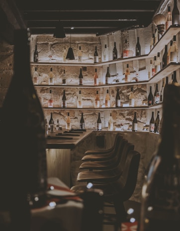 A dimly lit wine cellar with numerous wine bottles placed on wooden shelves that span across the stone walls. Bar stools with a rustic look are lined up alongside a wooden counter. The atmosphere is cozy and intimate, enhanced by the warm lighting that highlights the textures of the stone and wood.