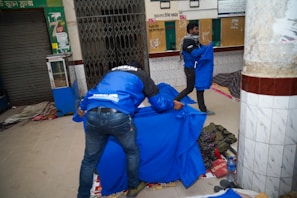 Two people are arranging vibrant blue covers over what appears to be sleeping areas on the ground. The location seems to be an indoor space with tiled walls and a closed storefront. Various items such as bags and a bottle are scattered around, suggesting a temporary or makeshift sleeping arrangement.