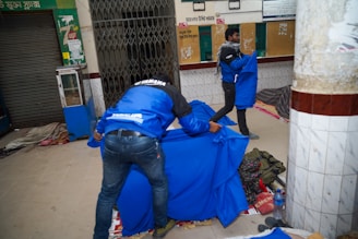 Two people are arranging vibrant blue covers over what appears to be sleeping areas on the ground. The location seems to be an indoor space with tiled walls and a closed storefront. Various items such as bags and a bottle are scattered around, suggesting a temporary or makeshift sleeping arrangement.