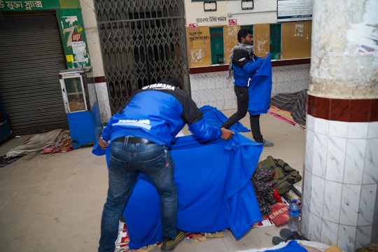 Two people are arranging vibrant blue covers over what appears to be sleeping areas on the ground. The location seems to be an indoor space with tiled walls and a closed storefront. Various items such as bags and a bottle are scattered around, suggesting a temporary or makeshift sleeping arrangement.