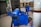 Two people are arranging vibrant blue covers over what appears to be sleeping areas on the ground. The location seems to be an indoor space with tiled walls and a closed storefront. Various items such as bags and a bottle are scattered around, suggesting a temporary or makeshift sleeping arrangement.