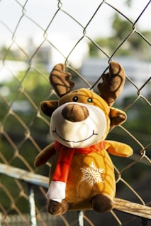 A close-up of a cheerful plush reindeer with big embroidered eyes and a red scarf, sitting on a wooden shelf.