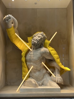 A classical stone sculpture of a muscular male figure is partially adorned with a modern bright yellow puffer jacket. The jacket is draped over the sculpture's back and shoulders, contrasting with the traditional art style of the sculpture. Wooden dowels are placed strategically in the display, interacting with the sculpture. The background is neutral, highlighting the artistic contrast between the old and new elements.