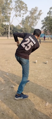 A person wearing a black shirt with the name 'Usman' and number '25' is playing cricket in an open field. The individual is preparing to bowl. The ground is dry with scattered leaves, and a few trees surround the area. Other people can be seen in the background enjoying the outdoor setting.