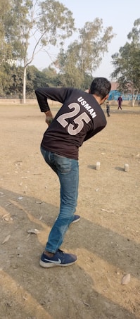 A person wearing a black shirt with the name 'Usman' and number '25' is playing cricket in an open field. The individual is preparing to bowl. The ground is dry with scattered leaves, and a few trees surround the area. Other people can be seen in the background enjoying the outdoor setting.