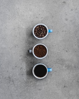 Three mugs are arranged vertically on a textured gray surface. The top mug, with a blue handle, contains whole coffee beans. The middle mug, with a black handle, is filled with ground coffee. The bottom mug, which also has a blue handle, is filled with brewed black coffee.