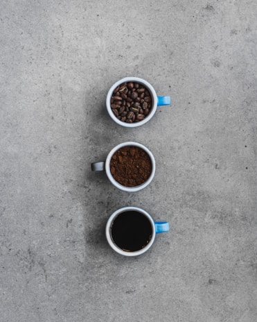 Three mugs are arranged vertically on a textured gray surface. The top mug, with a blue handle, contains whole coffee beans. The middle mug, with a black handle, is filled with ground coffee. The bottom mug, which also has a blue handle, is filled with brewed black coffee.